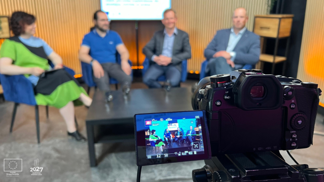 Priekšplānā TV kamera, fonā sēž četri cilvēki