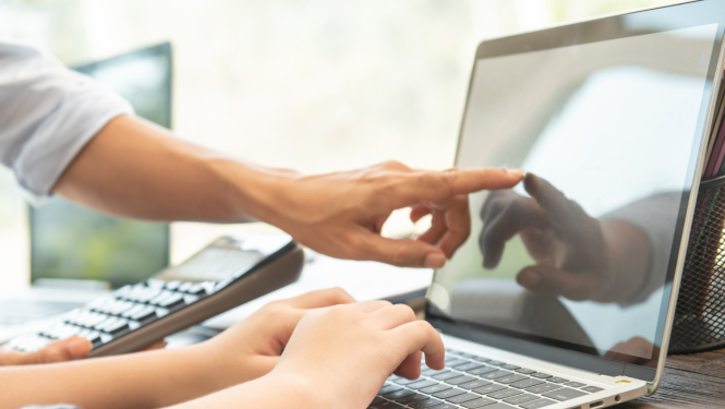 Cilvēka rokas uz klēpjdatora klaviatūras un rādītāja pirksts monitorā.