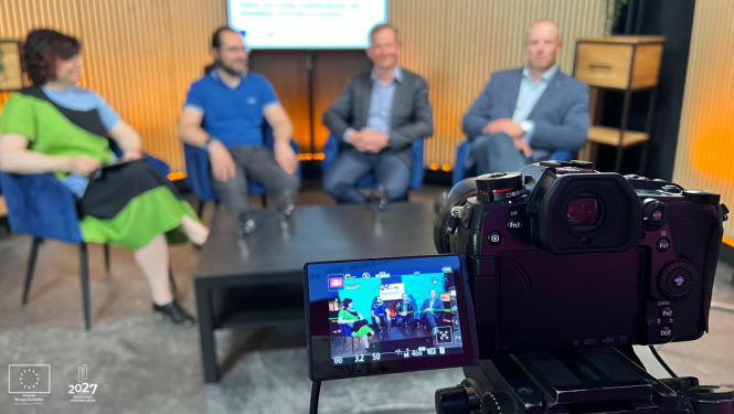 Priekšplānā TV kamera, fonā sēž četri cilvēki