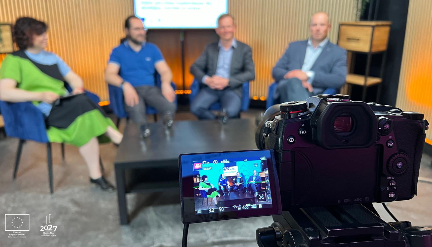 Priekšplānā TV kamera, fonā sēž četri cilvēki